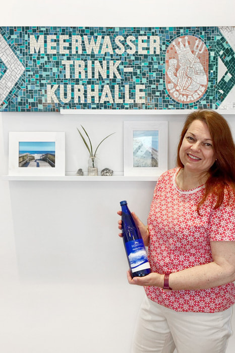 Shop-Betreiberin hält blaue BIOMARIS MeeresTIEFwasser Flasche vor Schild „Meerwasser Trink-Kurhalle“ , Bluse, Person, Portrait, Kräuter, Frau