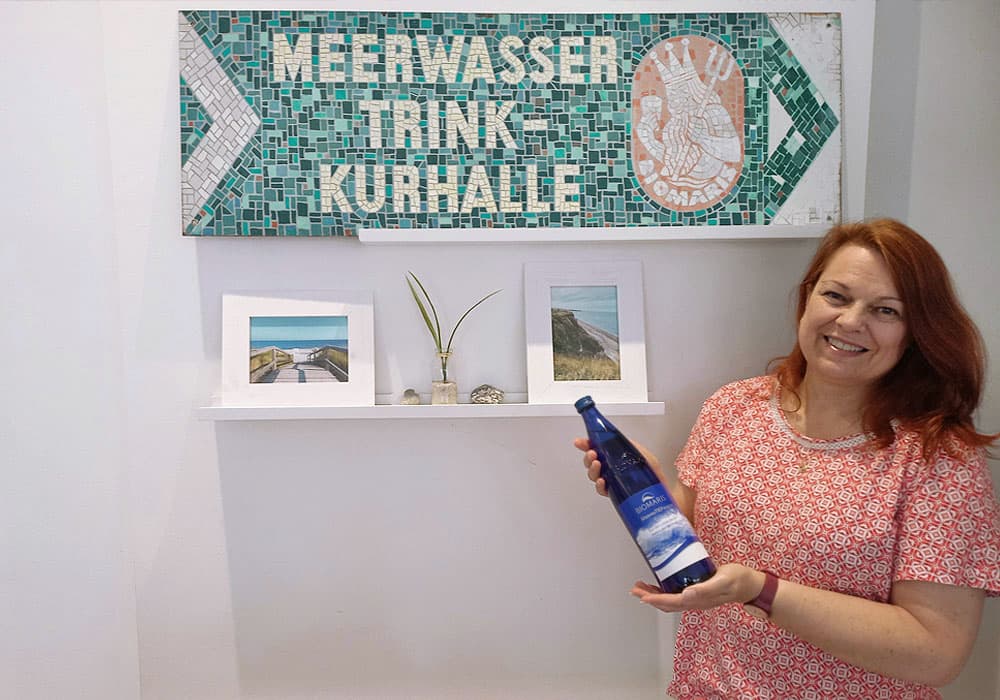 BIOMARIS Shop Heiligenhafen Shop-Betreiberin Shop-Betreiberin hält blaue BIOMARIS MeeresTIEFwasser Flasche vor Schild „Meerwasser Trink-Kurhalle“ , Bluse, Person, Portrait, Kräuter, Frau, Drinnen, Innenarchitektur, Werbung, Ärmel, Plakat