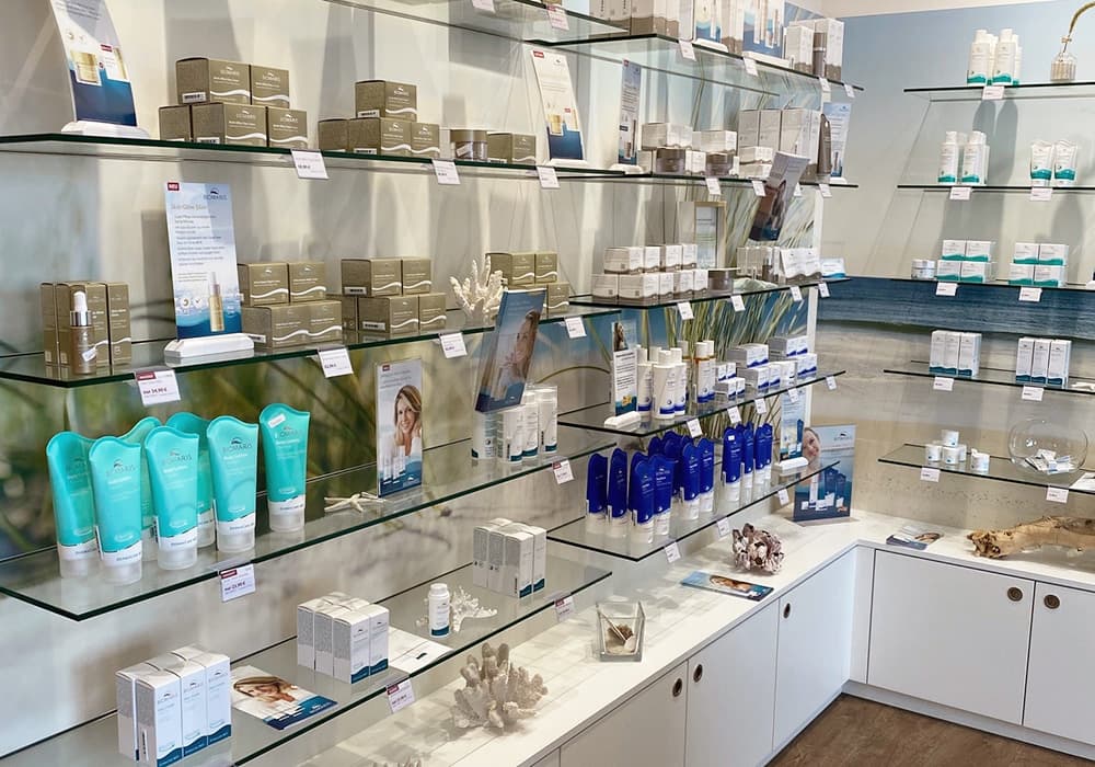BIOMARIS Shop Scharbeutz Wandregal mit BIOMARIS Pflegeprodukten auf Glasböden, darunter weiße Unterschränke auf Holzfußboden, Pflegeprodukt, Hautpflege, Körperpflege, Gesichtspflege, Feuchtigkeitscreme