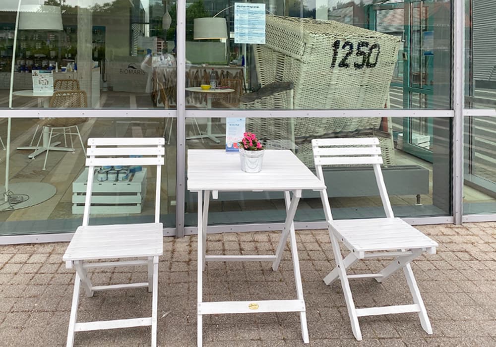 BIOMARIS Shop St. Peter-Ording Weiße Sitzgruppe mit zwei Klappstühlen und Tisch mit Blumentopf vor Glasfassade mit Strandkorb im Hintergrund